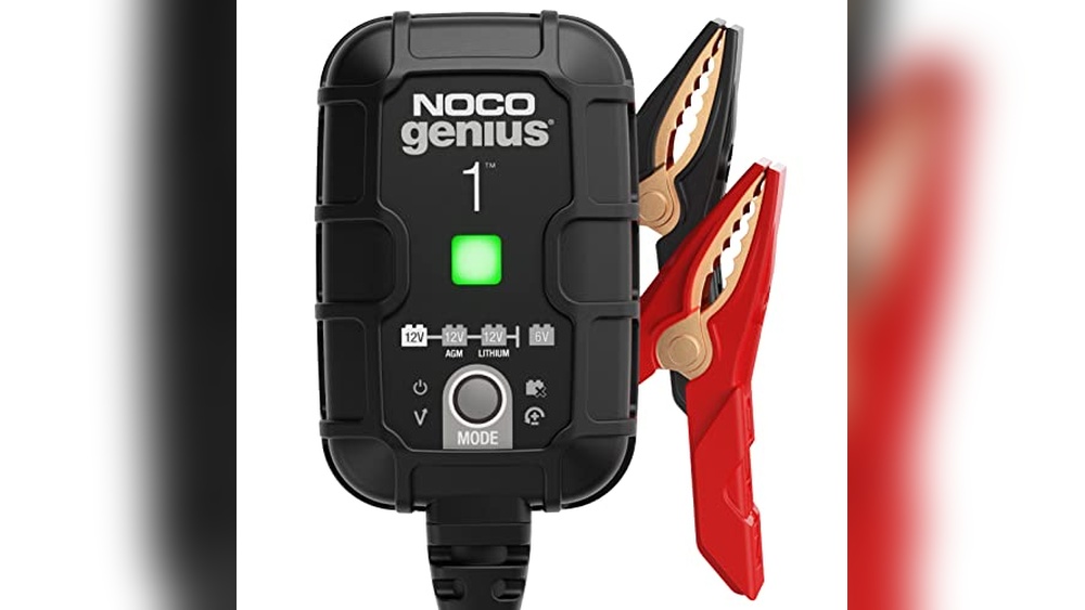 A NOCO Genius 1 battery charger with a green indicator light, attached black and red alligator clips, and labeled for 12V, AGM, and lithium batteries. The device is black with white text and icons.