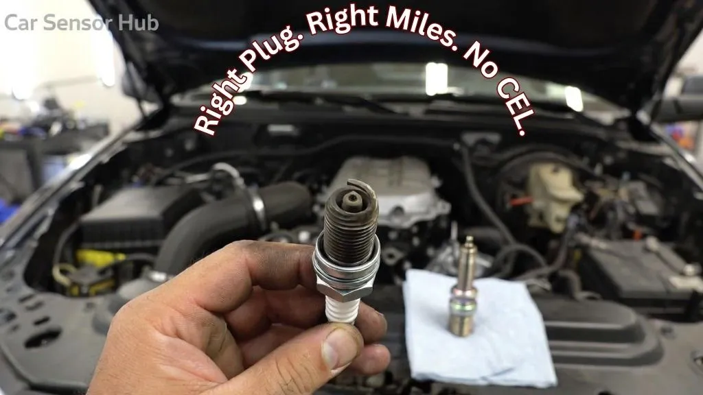 Mechanic holding a worn spark plug removed from a GMC Sierra 1500 engine bay next to a new iridium replacement plug on a shop rag
